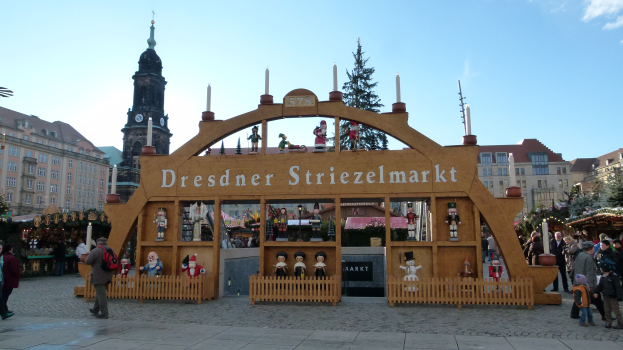 Ein lebendiger Weihnachtsmarkt in Dresden, Deutschland, mit Menschen, die herumlaufen, einem Bogen mit Text, Tannenbäumen, Gebäuden mit Fenstern und einer bewölkten Himmel.