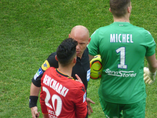Schiedsrichter spricht mit zwei Füßballspielern auf einem grünen Feld, ein Spieler hält einen Ball.