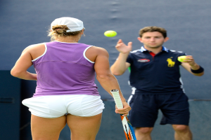 Eine Frau in einem lila T-Shirt, weißen Shorts und einer Mütze hält einen Schläger und schaut einem Mann in einem blauen Trikot zu, der einen Ball in ihre Richtung wirft, mit einem blauen Hintergrund.