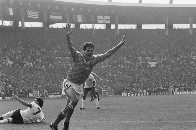 Ein Schwarz-Weiß-Foto eines Fußballspielers, der nach einem Tor jubelt, umgeben von Menschen auf dem Boden, mit Schildern, Fahnen und Pfosten im Hintergrund unter einem klaren Himmel.