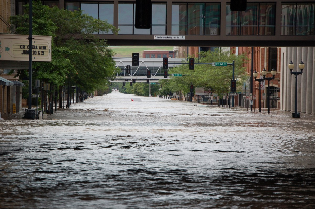 Überflutete Stadtstraße mit Wasser, das die Straße, die Infrastruktur und Gebäude bedeckt, einschließlich einer Brücke im Hintergrund.