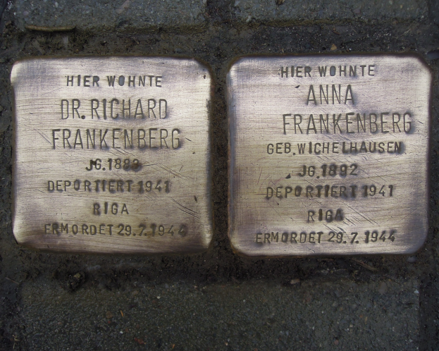 Zwei Messingplatten auf dem Boden vor einem Gebäude, mit Text beschriftet, der Holocaust-Opfer gedenkt.