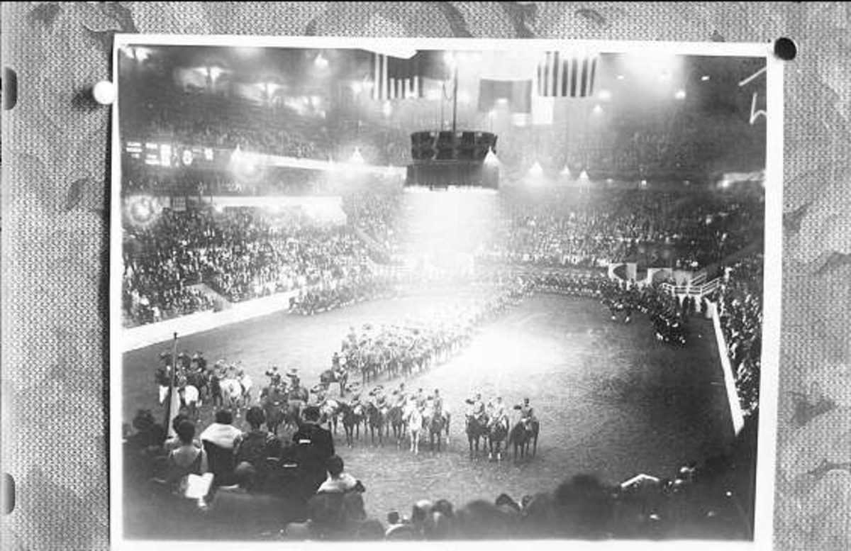 Schwarzes und weißes Foto einer großen Menschenmenge in einem Stadion, einige zu Pferd und andere stehend, mit einer Bühne in der Mitte und Text unten.