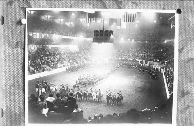 Schwarzes und weißes Foto einer großen Menschenmenge in einem Stadion, einige zu Pferd und andere stehend, mit einer Bühne in der Mitte und Text unten.