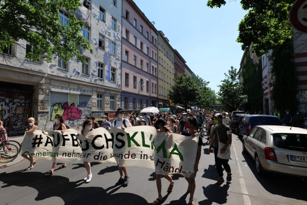 Eine Gruppe von Menschen marschiert bei einer Demonstration, hält ein Banner, mit Autos auf der rechten Seite, einem Radfahrer auf der linken Seite und Gebäuden, Bäumen und einem Schild im Hintergrund in Berlin.
