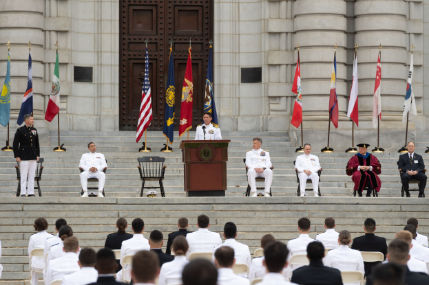 Gruppe von Menschen in weißen Uniformen auf Treppe eines Gebäudes mit Podium und Flaggen vor großem Gebäude mit Tür bei zeremoniellem Ereignis.
