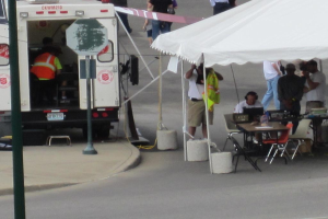 Ein Rettungswagen steht auf der linken Seite einer Straße, während auf der rechten Seite ein Zelt viele Menschen schützt, mit einem Tisch und Stuhl in der Nähe.
