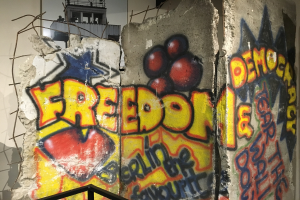 Mauer mit Graffiti, das "Freiheit und Demokratie" in fetten schwarzen Buchstaben liest, ein Geländer unten und ein Gebäude mit Fenstern im Hintergrund.