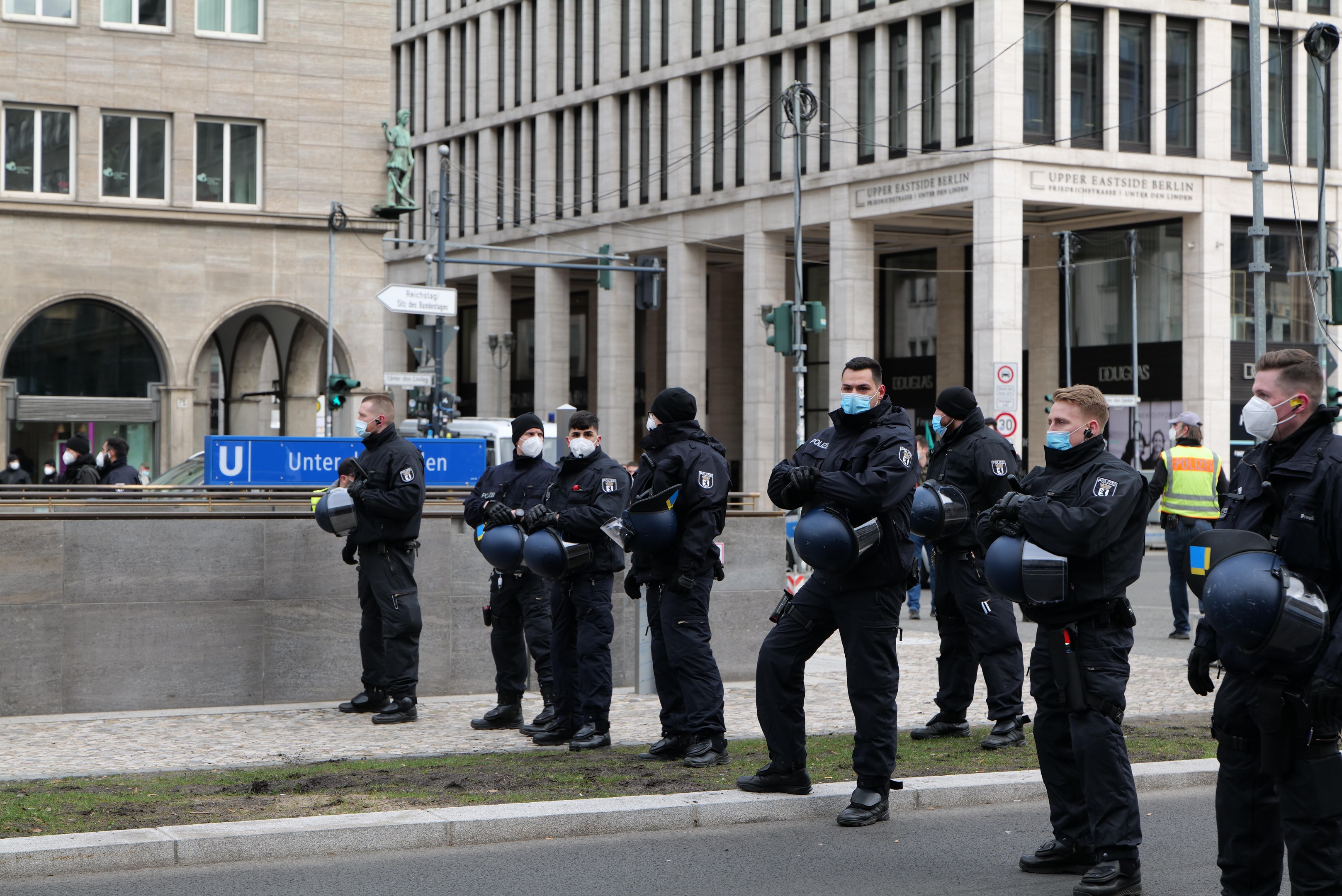 Eine Gruppe von Polizisten in schwarzen Uniformen und Masken steht vor einem Berliner Gebäude mit Glasfenstern, Säulen und grasbewachsenem Boden.