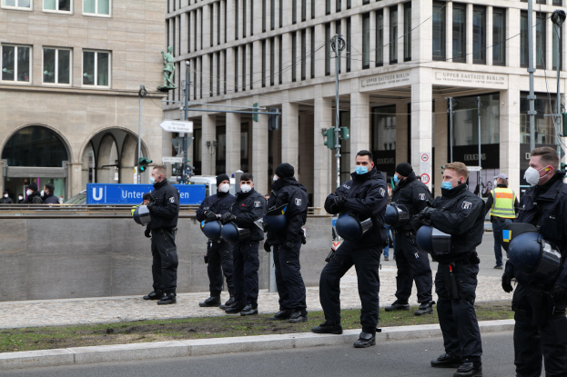Eine Gruppe von Polizisten in schwarzen Uniformen und Masken steht vor einem Berliner Gebäude mit Glasfenstern, Säulen und grasbewachsenem Boden.