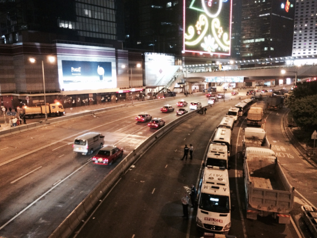 Eine belebte Stadtstraße bei Nacht mit hohen Gebäuden, Fahrzeugen, Passanten, Bäumen auf der rechten Seite und beleuchteten Plakaten im Hintergrund.