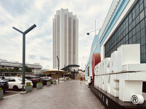 Ein belebter zentraler Markt in Kuala Lumpur, Malaysia, mit hohen Gebäuden, Straßenmasten, Straßenlaternen, Kraftfahrzeugen, Fußgängern auf dem Gehweg, Namensschildern, Bäumen und einem bewölkten Himmel.