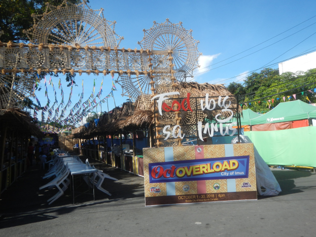 Eine Karnevals-Szene mit einem großen Riesenrad in der Mitte, umgeben von Hütten, Sitzbereichen, einer Tafel, Dekorationen, Bäumen, Überlandleitungen und einem bewölkten Himmel.