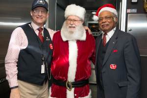 Eine Gruppe von Menschen mit Brillen, Mützen, Krawatten, ID-Karten und Ausweisen, wobei eine Person ein Weihnachtskostüm trägt, vor einer Stahlwand mit Lichtern, Schildern und anderen Gegenständen im Hintergrund steht.