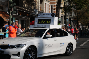 Ein weißer BMW fährt die Straße entlang, mit einer Uhr auf dem Dach, während eine Person im Auto sitzt und Menschen auf der Straße stehen. Im Hintergrund sind Gebäude, Bäume und Ampeln zu sehen.