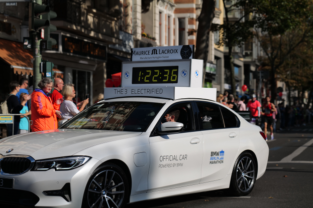 Ein weißer BMW fährt die Straße entlang, mit einer Uhr auf dem Dach, während eine Person im Auto sitzt und Menschen auf der Straße stehen. Im Hintergrund sind Gebäude, Bäume und Ampeln zu sehen.