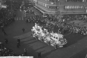 Schwarzes und weißes Foto einer festlichen Parade mit einem dekorierten Wagen, umgeben von einer großen Menge in bunter Kleidung, vor Gebäuden mit Fenstern und Laternenmasten.