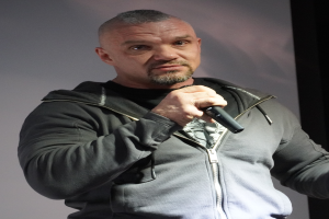 Ein Mann in schwarzer Kapuzenjacke, schwarzem Anzug und wei├čem Hemd mit zurückgek├Ąmmten Haaren h├Ąlt ein Mikrofon und steht selbstbewusst vor einer Wand.