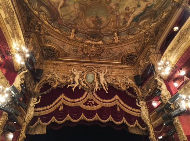 Das Innere eines Theaters mit einem roten Vorhang, Kronleuchtern, Skulpturen, Lichtern und einer bemalten Decke, mit einer Bühne und ein paar Menschen unten.