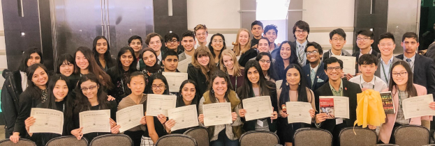 Gruppe von Menschen mit Urkunden, wahrscheinlich Absolventen des akademischen Jahres 2019-2020, die für ein Foto in einem Raum mit Stühlen und einer Wand im Hintergrund posieren.