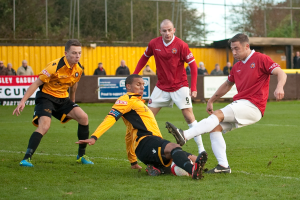 Spieler in blauen und roten Uniformen spielen ein Spiel auf einem Rasenfeld mit einem Ball, während Zuschauer außerhalb des Spielfelds stehen und sie anfeuern, mit Bäumen und Himmel im Hintergrund.
