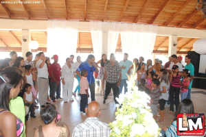 Eine Gruppe von Menschen mit einer Pflanze und einem Logo unten, Vorhänge und ein Lautsprecher im Hintergrund und Text in der oberen linken Ecke.