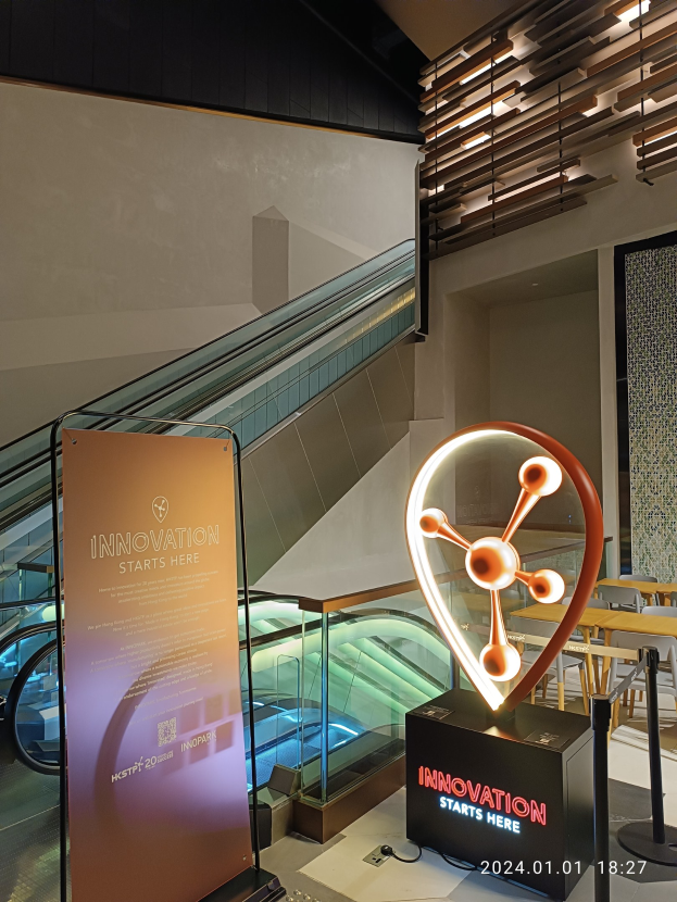 Großes Schild mit der Aufschrift "HSTP20 Expo" in einem Gebäude mit einer Treppe im Hintergrund, Tische und Stühle auf der rechten Seite und eine Tafel mit "Innovation beginnt hier" auf der linken Seite.