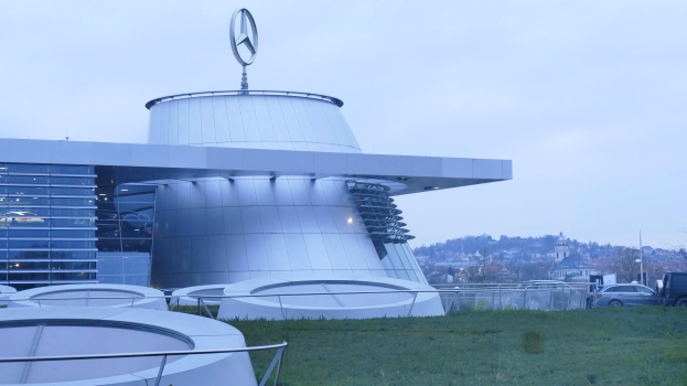 Das Mercedes-Benz Museum in Frankfurt, Deutschland, ein großes Gebäude mit einem Logo darauf, umgeben von Gras, Geländern, Fahrzeugen auf der Straße, Bäumen und einem klaren blauen Himmel.
