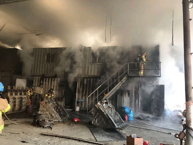 Feuerwehrleute in Helmen verwenden Schläuche, um ein Lagerfeuer zu löschen, mit Rauch, der aus dem Gebäude aufsteigt, Treppenstufen mit Geländern, die zum Eingang führen, einem Kettenglied an der rechten Seite, einer fensterumrandeten Wand im Hintergrund und verstreuten Trümmern, einschließlich Kartons und Rohre auf dem Boden.