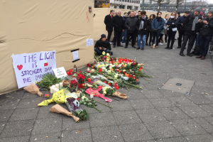 Eine Gruppe von Menschen steht vor einem Zelt mit einer Tafel, auf der 'Das Licht ist stärker als die Dunkelheit' steht, umgeben von Blumen, Kerzen und anderen Huldigungen, mit Gebäuden und Bäumen im Hintergrund.