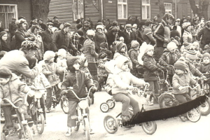 Eine Gruppe von Kindern fährt auf Fahrrädern eine Straße entlang vor einer Menge von Menschen, einige tragen Käpchen, mit Bäumen und einem Fenster mit Fenstern im Hintergrund, dargestellt in Schwarz-Weiß.