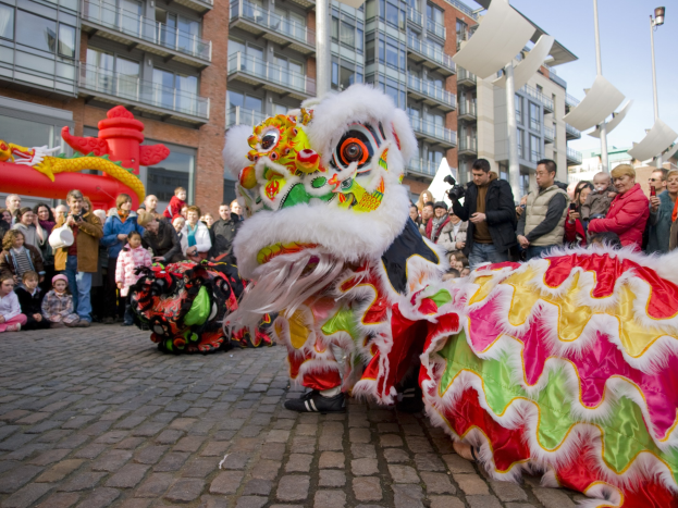 Ein lebendiges chinesisches Neujahrsfest in Amsterdam mit einem Drachen tanzen und einer Menge Schaulustiger, einige fotografieren, vor einem Hintergrund von Gebäuden, Laternenmästen und einem klaren blauen Himmel.
