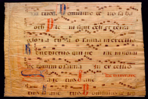 Ein illuminiertes Manuskript mit Musiknotation auf einem Blatt Papier vor dunklem Hintergrund, gefüllt mit Text und Musiksymbolen, die ein komplexes Design bilden.