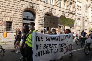 Eine Gruppe von Menschen marschiert auf einer Straße in Berlin, hält Schilder und Banner hoch und einige fahren Fahrräder, vor einem Gebäude mit Fenstern, Bögen, Säulen, Skulpturen, Bäumen und einem Laternenmast, an einer Anti-Kriegs-Protestaktion teilnehmend.