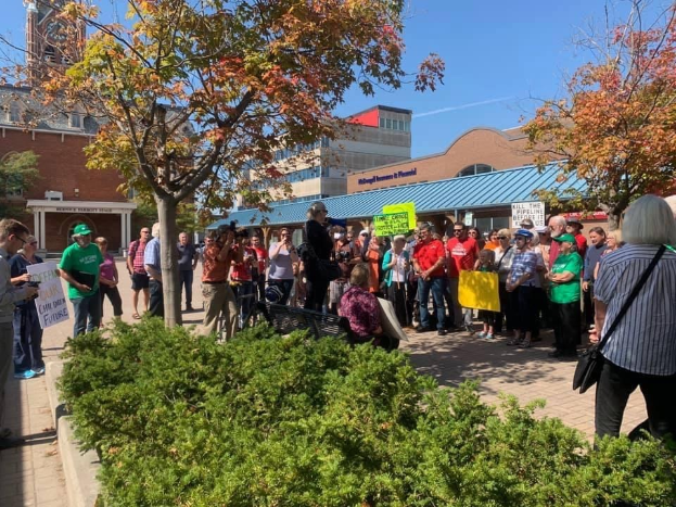 Eine Gruppe von Studenten hält Protestschilder vor einem Gebäude auf dem Campus der University of Michigan, mit Bäumen und Himmel im Hintergrund.
