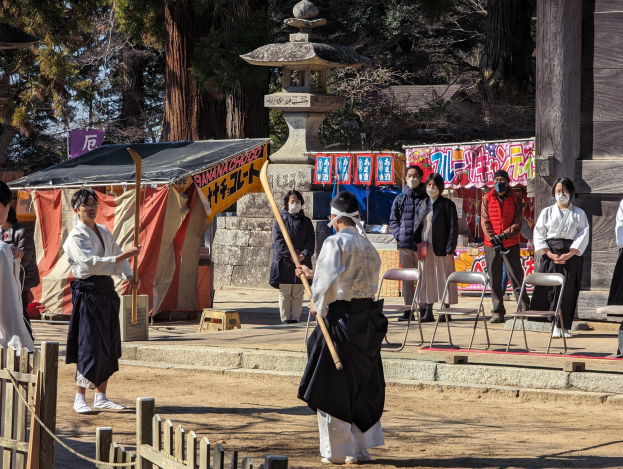 Eine Gruppe von Menschen in formeller Kleidung, einige mit Masken und Holzstöcken, versammelt sich im Freien vor einem traditionellen Gebäude während einer Zeremonie in Kyoto, mit Stühlen, Bannern, einem Zelt und Bäumen unter einem klaren blauen Himmel.