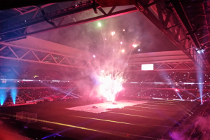 Ein großes Stadion voller Menschen, die ein Feuerwerk bewundern, mit einer hell erleuchteten Bühne mit Trägern und Scheinwerfern oben und einem Netz auf der linken Seite.