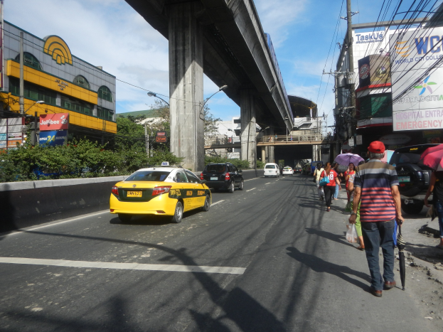Gelbes Taxi fährt auf einer Straße mit hohen Gebäuden, Menschen mit Schirmen auf dem Gehweg, eine Brücke im Hintergrund, bewölkter Himmel und Strommasten mit Drähten entlang der Straße.