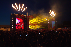 Eine große Menschenmenge auf einem Musikfestival schaut Performern auf einer hell erleuchteten Bühne zu, mit Zelten links und Feuerwerk am dunklen Himmel dahinter.