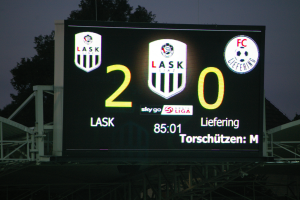 Ein großes digitales Anzeigebrett, das das Ergebnis eines Fußballspiels und Logos anzeigt, vor einer Kulisse aus einem Stadion, Bäumen und einem klaren blauen Himmel.
