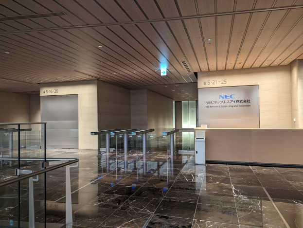 Lobby eines Gebäudes mit Marmorboden, Glasgeländern, elektrischen Lampen, die bis zur Decke reichen, Namensschildern an den Wänden und einem Empfangstisch mit dem Logo von NEC Network & System Integration Corporation.