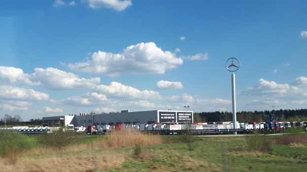 Eine Mercedes-Benz-Fabrik in Deutschland mit Fahrzeugen auf der Straße, umgeben von Grünflächen und Gebäuden, unter einem bewölkten Himmel.