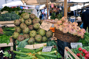Körbe gefüllt mit Obst, Gemüse und kleinen Schildern mit Text, mit Zelten und einigen Menschen im Hintergrund.