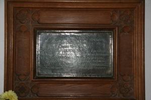 Eine Gedenktafel mit Text ist an einer braunen Holztür oder Wand angebracht, mit einem gelben Gegenstand, der einer Vase ähnelt, in der linken unteren Ecke.