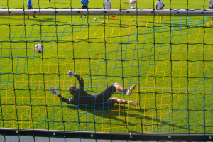 Ein Fussballspieler hält den Ball vor einem Tor, mit einem Netz im Vordergrund und einigen Menschen im Hintergrund sowie mehreren Bällen auf dem Boden verteilt.