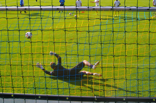 Ein Fussballspieler hält den Ball vor einem Tor, mit einem Netz im Vordergrund und einigen Menschen im Hintergrund sowie mehreren Bällen auf dem Boden verteilt.