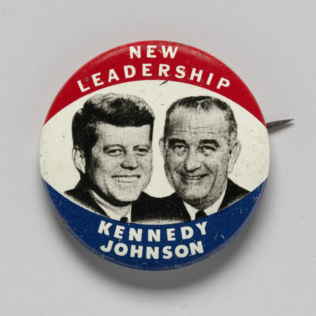 Weißer Ansteckbutton auf einer Oberfläche, der zwei Fotos von Kennedy und Johnson mit dem Text "Neue Führung" darunter zeigt.