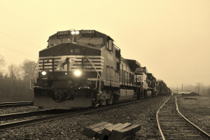 Ein Zug auf Eisenbahnschienen mit Autos, Strommasten, Bäumen und einem Himmel im Hintergrund.