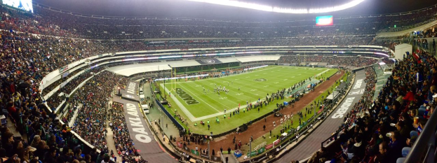 Ein großes Stadion voller Zuschauer, die ein Fußballspiel sehen, mit Spielern und verschiedenen Gegenständen auf dem Feld, beleuchtet von Deckenlampen, und mit Tafeln, Geländern und Bildschirmen.