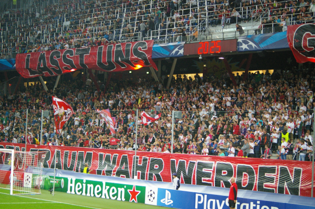 Fussballspiel in einem Stadion mit einer großen Menge, grünem Rasen, Torpfosten links, Bodenbanner, Menschen mit Fahnen und Hintergrundgeländer, Tafeln, Lichter und andere Gegenstände.